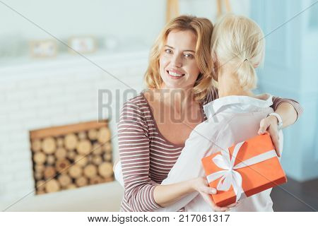 Loving granddaughter. Pretty cheerful calm woman holding a beautiful present in her hands while feeling happy and hugging her kind grandmother