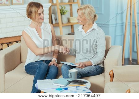 Deal. Responsible experienced young financial advisor looking happy while shaking hands with her cheerful pleasant aged client