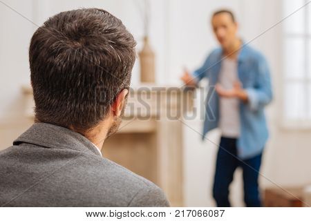 Severe problem. Experienced focused male psychologist turning to the patient who standing while positing on the blurred background