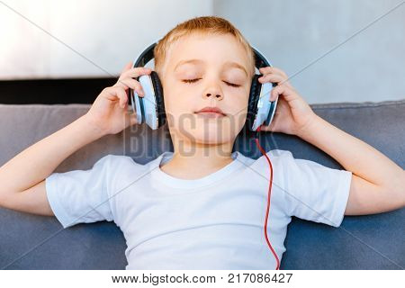 Pleasurable rest. Pleasant nice calm boy holding his headphones and listening to music while feeling relaxed