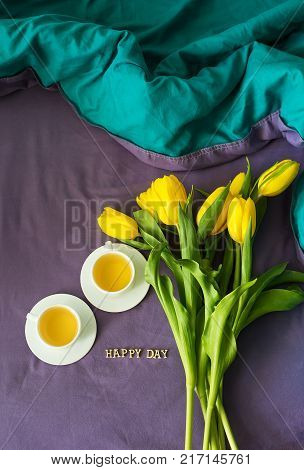 Yellow tulips and two cups of tea in bed, close-up, lettering happy day