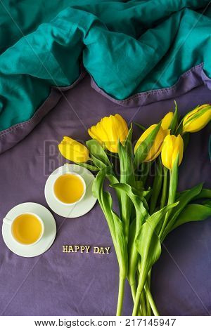 Yellow tulips and two cups of tea in bed, close-up, lettering happy day