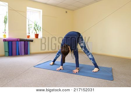 Sportswoman focused on stretch before Pilates class. Girl doing warm up for legs on mat. Female has hair pulled back, concentrated look. Athlete dressed in dark top, yoga legging trains in fitness room. Concept of online training, customizable workouts.