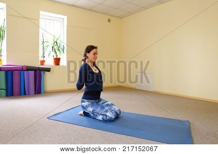 Sportswoman focused on stretch before Pilates class. Girl doing warm up for legs on mat. Female has hair pulled back, concentrated look. Athlete dressed in dark top, yoga legging trains in fitness room. Concept of online training, customizable workouts.
