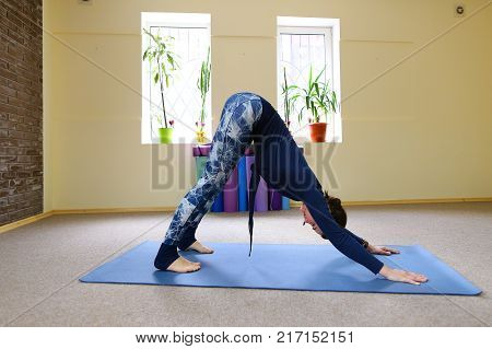 Sportswoman focused on stretch before Pilates class. Girl doing warm up for legs on mat. Female has hair pulled back, concentrated look. Athlete dressed in dark top, yoga legging trains in fitness room. Concept of online training, customizable workouts.