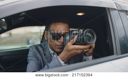 African american paparazzi man sitting inside car and photographing with slr camera