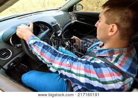 Handsome serious young man in plaid shirt driving his car and using smart phone. Young man with hand on steering wheel using mobile phone while driving. Doing forbidden things