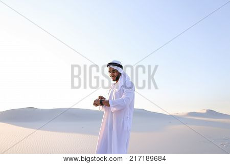 Attractive guy, emirate businessman uses for business and for work Watch, who are put on hand, smiling and examining landscapes of large sandy desert against blue sky on hot summer morning. Swarthy Muslim with short dark hair dressed in kandura