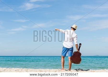 Back view of man holding guitar and standing on the beach with blue sea and blue sky bacjground. Travel relax vacation concept.