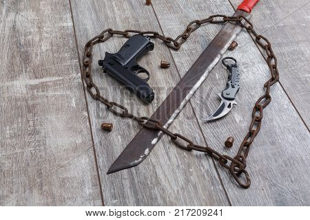 A rusty chain in the shape of a heart and a machete that pierces the heart, a pistol and a knife on a wooden background. Top view from an angle