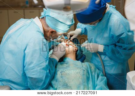 Male Old Professional Dentist Surgeon In Uniform And Female Assistant Helps To Performing Operation