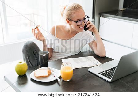 Beuatiful concentrated lady working with laptop and talking on smartphone while eating breakfast in morning