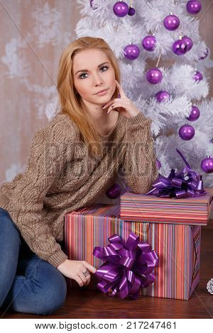 Amazing beautiful face lady girl with perfect skin and blond hairs posing for christmas holidays close to new year white pine tree and presents in studio.