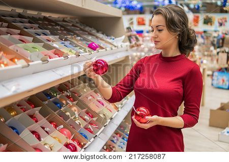 Cheerful happy customer girl choosing gifts for Christmas and New Year. She is taking red balls Christmas tree decorations. difficult decision. Various choice. She is smiling