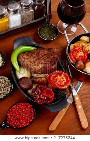 fresh roast beef meat rib eye steak with tomatoes,potatoes, hot chili pepper gern and pink peppercorn saucer, cutlery, set of spice, and red wine glass on light walnut table