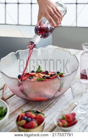 Pouring red wine into bowl filled with sliced fruit strawberries blueberries and fresh herbs, preparing sangria party punch