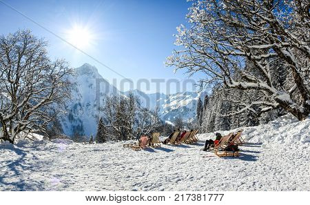 Group of people sitting with deck chairs in sunny winter mountains. Sunbathing in snow. Germany, Bavaria, Allgau, Schwarzenberghuette.