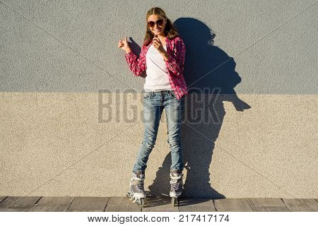 Portrait of young cool girl, shod in rollerblades holding chocolate bar and showing thumbs up. Thumb up energy and joy