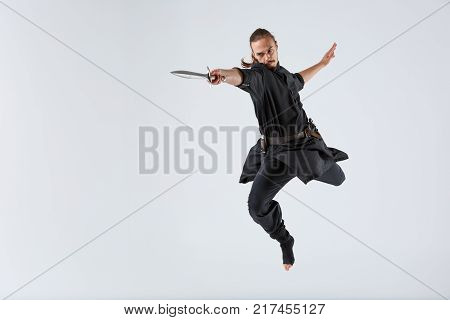 A ninja man in black uniforms in a jump with a sword in his outstretched hand and a hand stretched out back. Front view on a gray background
