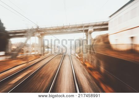 View from cabin of high-speed suburban train on autumn evening: railway tracks going to vanishing point autumn grass bridge pillars with wires station and buildings; strong motion blur