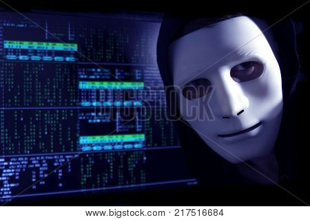 A hacker in a mask on a background of binary code looks into the frame.
