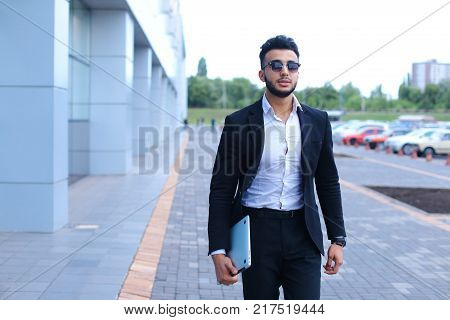 Young handsome businessman Arab Muslims is in the business center in black elegant dress on building background. Businessman man male looking at time walking goes and smiling.