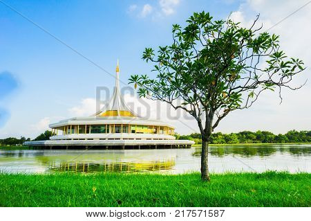 Thai style auditorium is middle of the water. This auditorium is building oval and paint yellow color in building. It have grass and tree in foreground forest in background on blue sky day.