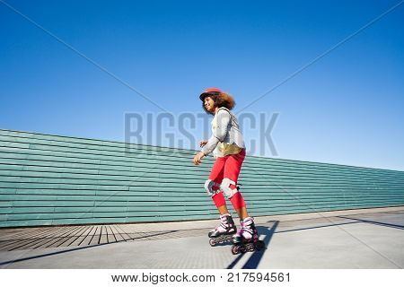 Side view portrait of African girl in helmet and protective gear, riding fast at rollerdrom against blue sky