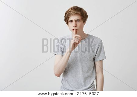Portrait of angry irritated fair-haired young male asks to keep confidential information in secret, annoyed with gossiping people, demands quiet, says: Hush, stop talking. Caucasian guy gestures, asks for silence
