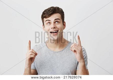 Portrait of young caucasian guy wearing gray t-shirt pointing at copy space up with his forefingers looking with broad smile upwards demonstrating something at white studio background. Body language concept