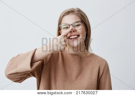 Gloomy young caucasian female makes fun, touches her nose, mocks, laughs and broadly smiles, demonstrates her white even teeth. Positive blonde girl joyfully smiles at camera