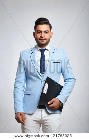 Studio portrait of a handsome bearded young man dresses in bkue jacket and white trousers posing with a clipboard against light grey background stylish style fashion hipster concept.