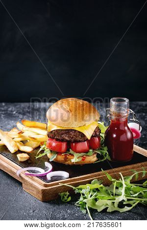 Homemade traditional hamburger with beef, tomato, cheese, aragula, served on wooden slate serving board with french fries and ketchup sauce on dark texture background.
