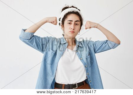 Brunette female wearing do-rag and denim shirt, shows muscles and strength, has confident expression, ready to fight or protect herself. Cute tender woman has power, being sure in victory.