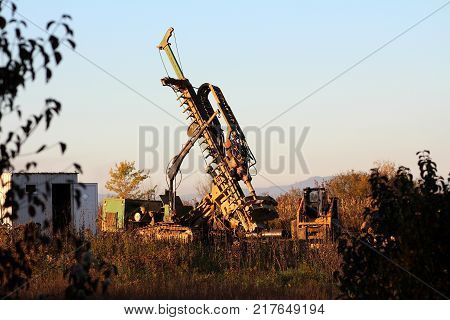 Portable trailer mounted industrial water boring machine rig with accompanying air compressor, metal pipes, small digger, large metal container for workers with open doors surrounded with high uncut grass and other vegetation at sunset