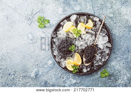 Black caviar in a glass bowl on ice with lemon close up over gray background, top view
