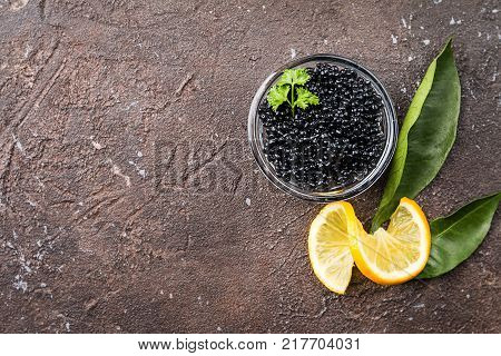 Black caviar in a glass bowl with lemon close up over dark background, top view
