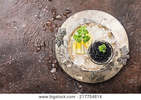 Black caviar in a glass bowl on ice with lemon close up over dark background, top view