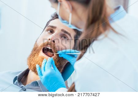 Side view of a reliable female dentist, using sterile instruments and blue surgical gloves while cleaning the teeth of a patient in the dental office of a modern clinic