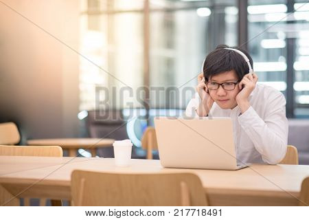 Young Asian man dressed in casual style listening to music while working with his laptop computer. Digital nomad in co working space modern IT lifestyle with work life balance concept.