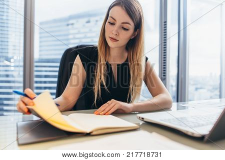 Confident female entrepreneur planning her workday sitting at her desk holding pen thinking looking at notebook in office.