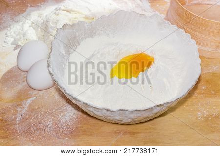Bowl with sifted wheat flour and broken egg in it and two whole eggs kneaded dough and wooden sieve beside on the table