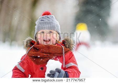 Little boy playing with snowballs with snowman on background. Active outdoors leisure with children in winter. Kid during stroll in a snowy winter park. Snowball fight