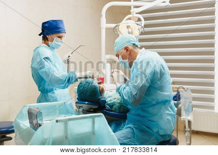 Male Old Professional Dentist Surgeon In Uniform And Female Assistant Helps To Performing Operation