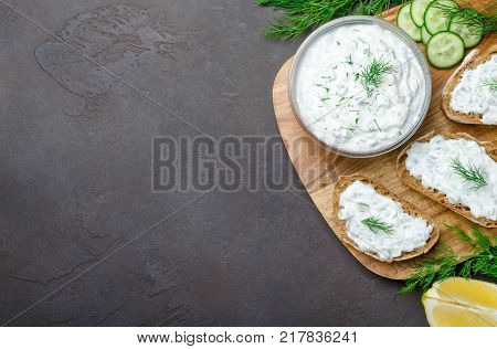 Homemade greek tzatziki sauce in a glass bowl with ingredients and sliced bread on a dark black stone background. Cucumber, lemon, dill, garlic. Top view, horizontal image, copy space