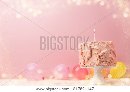 Birthday cake with pink frosting and burning single candle in arrangement with balloons in sparks.