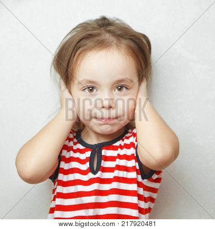 Adorable little girl with sincere smile and big brown eyes covers her ears with palms dressed in striped T-shrit with anchor stands beside white wall portrait photo.