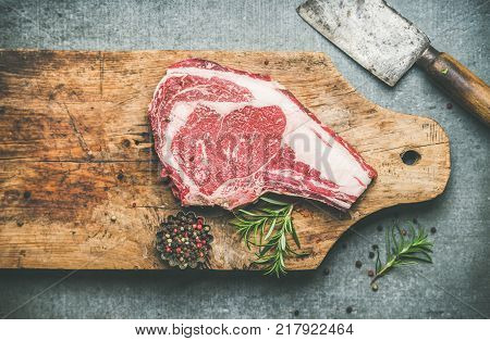 Flat-lay of raw prime beef meat dry-aged steak rib-eye on bone with seasoning and chopper knife on wooden board over grey concrete background, top view, copy space. Meat high-protein dinner concept