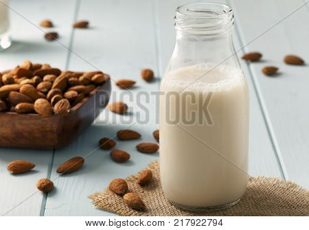 Almond Milk in glass bottle and tumbler with almond nuts in a wooden bowl on light blue table top with fringed Hessian coaster mat early morning sunlight from left.