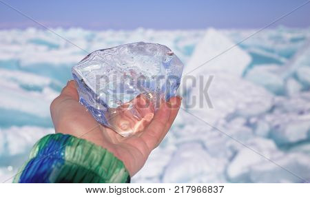 A piece of crystal transparent ice in the hand against frozen lake in sunny day